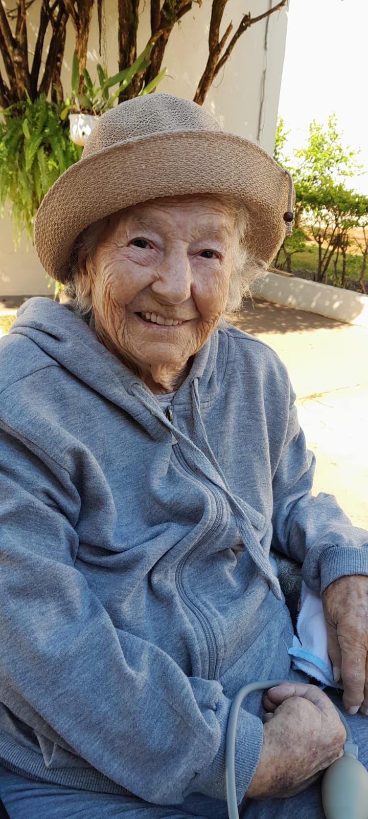 Senhora idosa sorrindo, usando chapéu bege e blusa azul, sentada ao ar livre